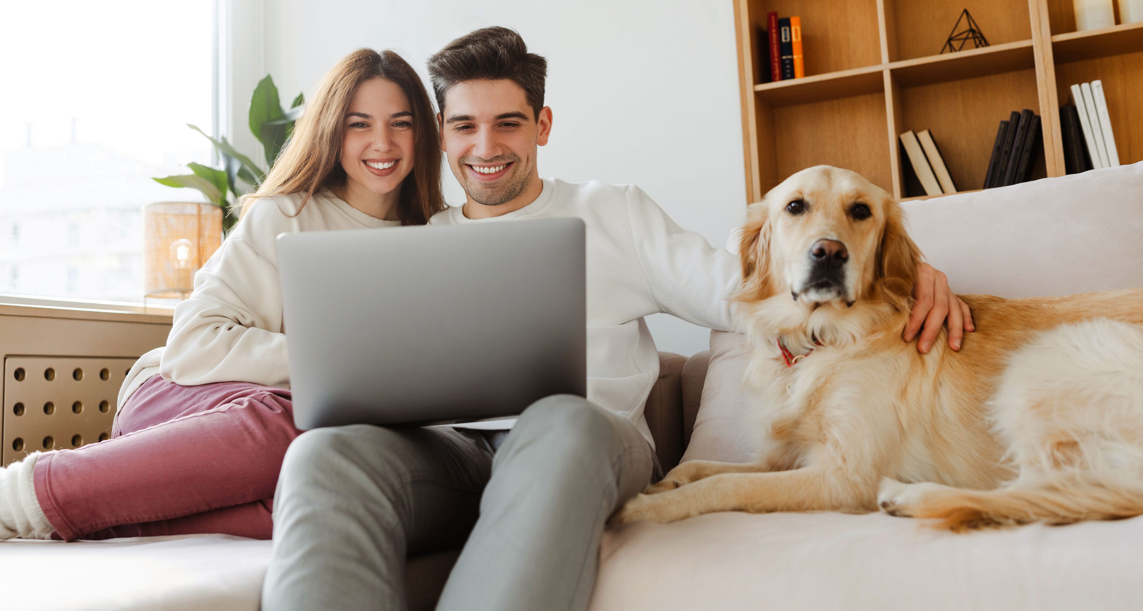 Happy young couple using laptop computer shopping online, watching video sitting with dog on comfortable sofa. Smiling man and woman working from home